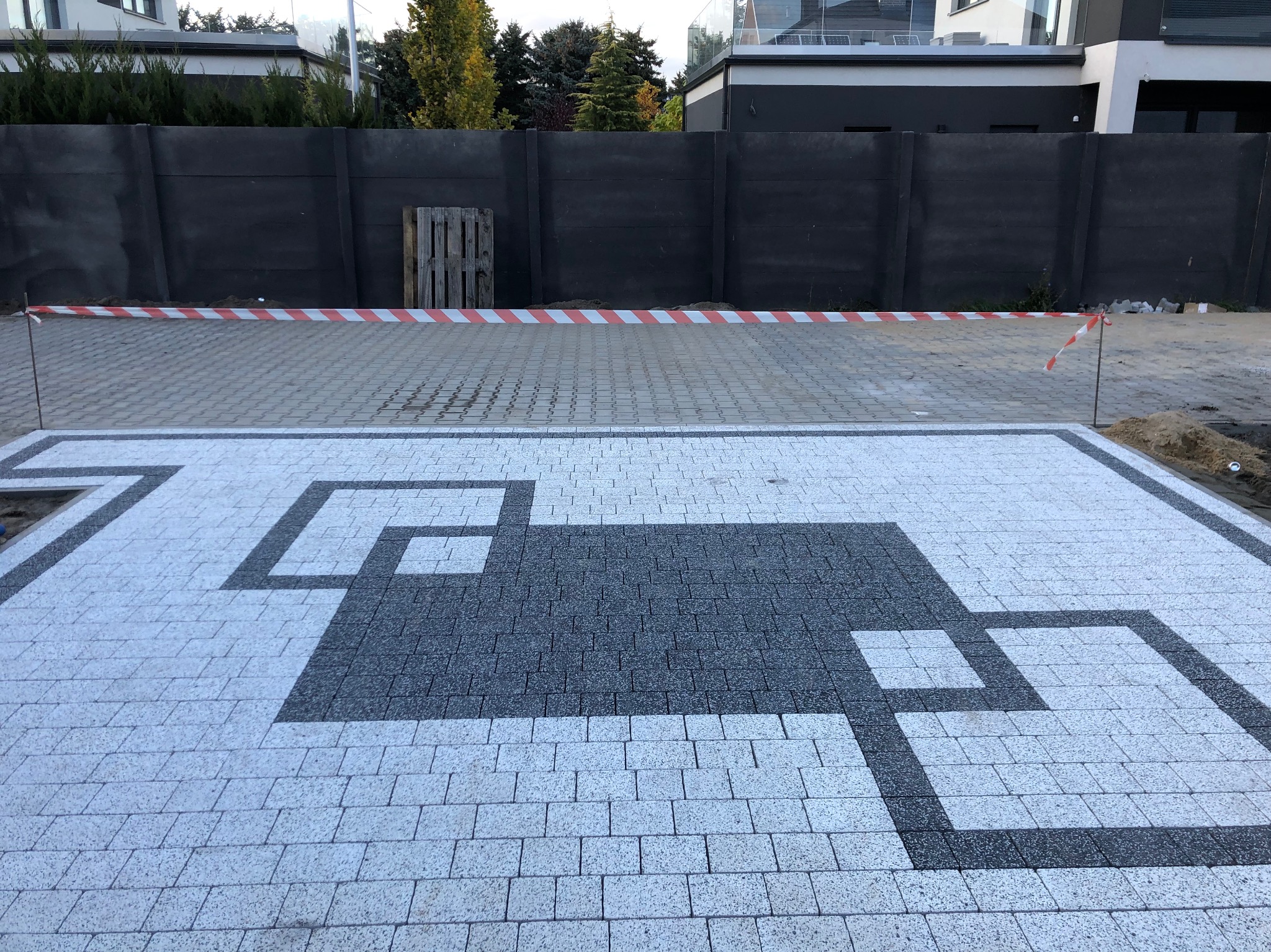 Ułożona kostka brukowa w nowoczesnym wzorze geometrycznym z szarych i białych kostek, z widoczną taśmą ostrzegawczą i paletą w tle, na tle ciemnego ogrodzenia i domu.