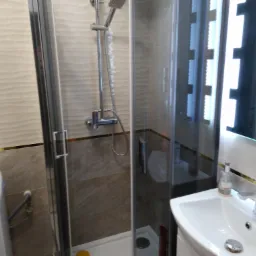 Nowoczesna kabina prysznicowa z deszczownicą i baterią termostatyczną, umywalka z armaturą i dozownikami mydła, pralka w tle, wykończenie w odcieniach beżu z dekoracyjnym paskiem