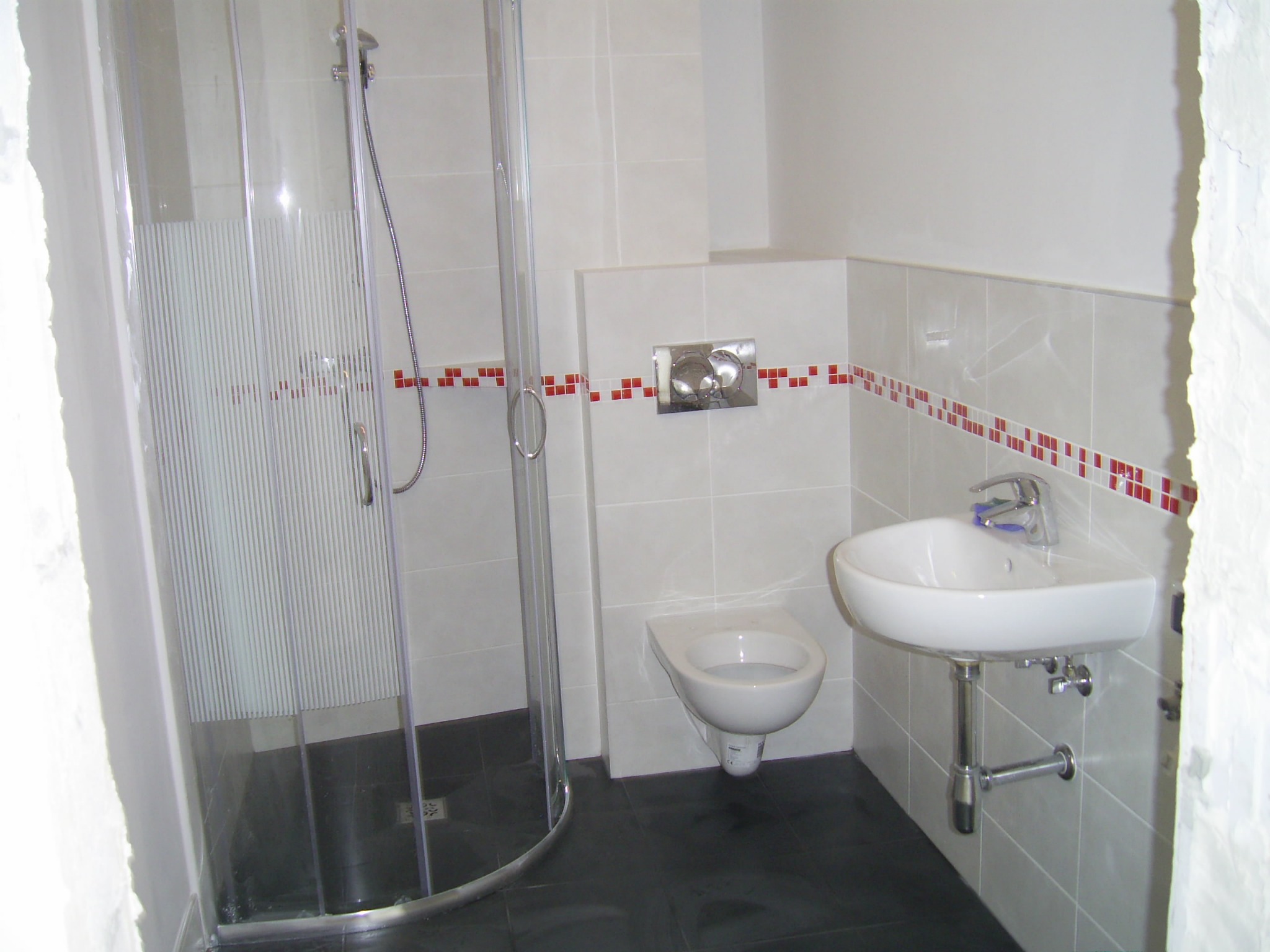 Nowoczesna łazienka z kabiną prysznicową z transparentnego szkła, podwieszaną toaletą i białą umywalką na tle białych płytek z dekoracyjnym pasem czerwonych kwadratów.