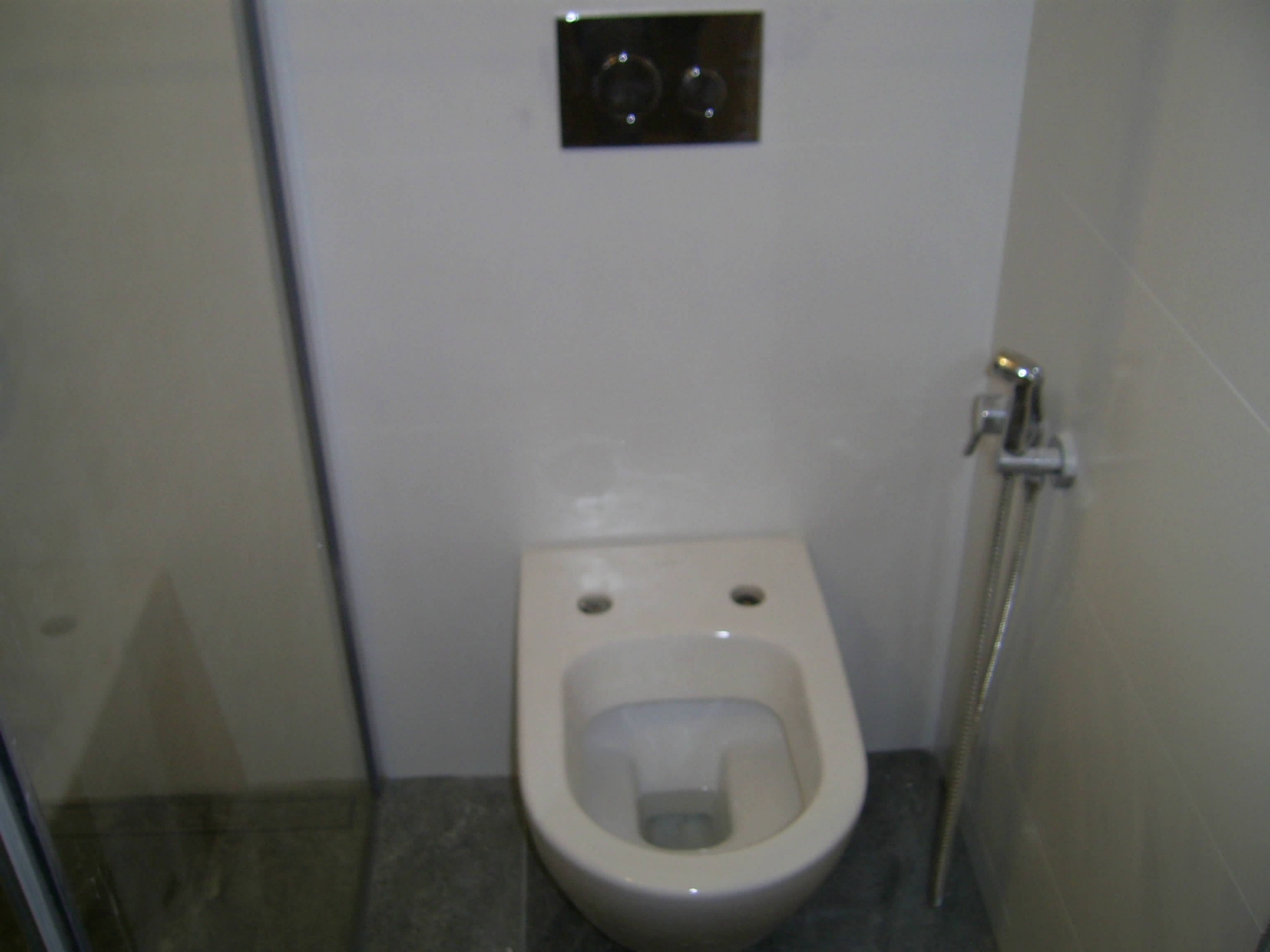 Wisząca toaleta z chromowanym panelem spłukującym i bidetką w jasnej łazience z szarymi płytkami podłogowymi.