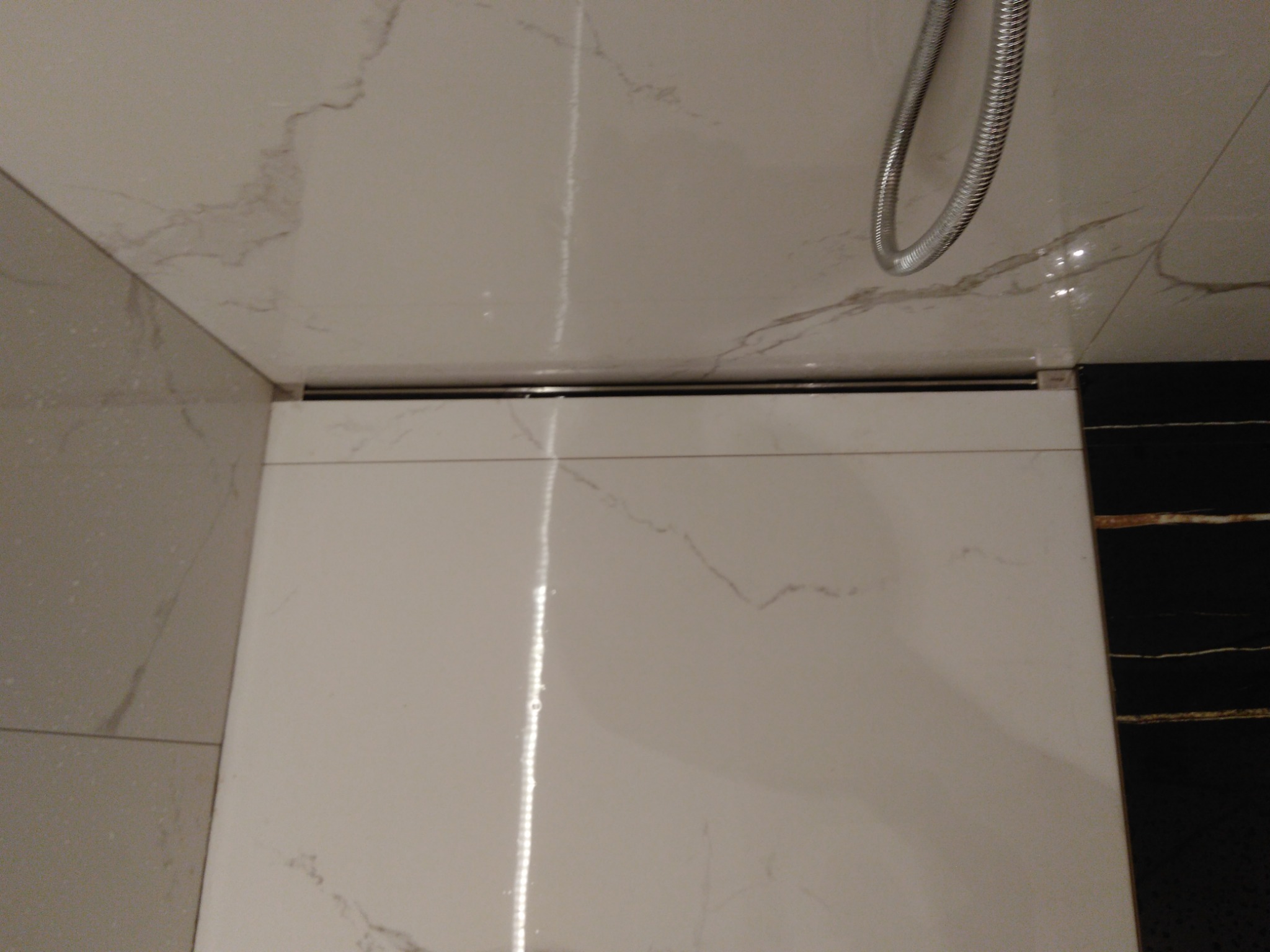 Szczegółowe ujęcie odpływu liniowego w kabinie prysznicowej wyłożonej białymi płytkami imitującymi marmur, z widocznym fragmentem węża prysznicowego i czarną ścianą w tle.