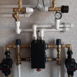 Rozbudowany węzeł hydrauliczny z czarną skrzynką rozdzielczą, manometrem i dwiema pompami obiegowymi Grundfos zamontowany na ścianie o jasnej fakturze.