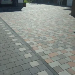 Ułożona kostka brukowa w dwóch odcieniach szarości i beżu, oddzielona ciemną linią, tworząca wzór na podjeździe przed garażem i budynkiem gospodarczym w słoneczny dzień.