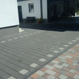 Ułożona kostka brukowa w dwóch odcieniach szarości i czerwieni, prowadząca do białego budynku z garażem i ławką przed wejściem.