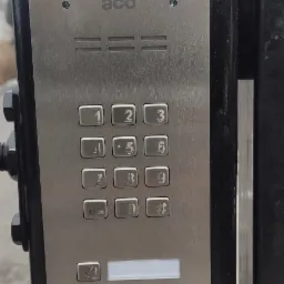 Metalowy panel domofonu z klawiaturą numeryczną i przyciskiem dzwonka, zamontowany na czarnej, metalowej ramie przy wejściu.