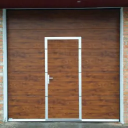 Segmentowa brama garażowa z drzwiami przejściowymi w kolorze drewna, z jasną ramą, zamontowana w murze z czerwonej cegły.