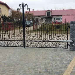 Otwarta, dwuskrzydłowa brama wjazdowa z czarnego metalu o dekoracyjnych, roślinnych ornamentach, zamontowana na słupkach z szarej kostki, z widocznym napędem ramieniowym i kostką brukową...