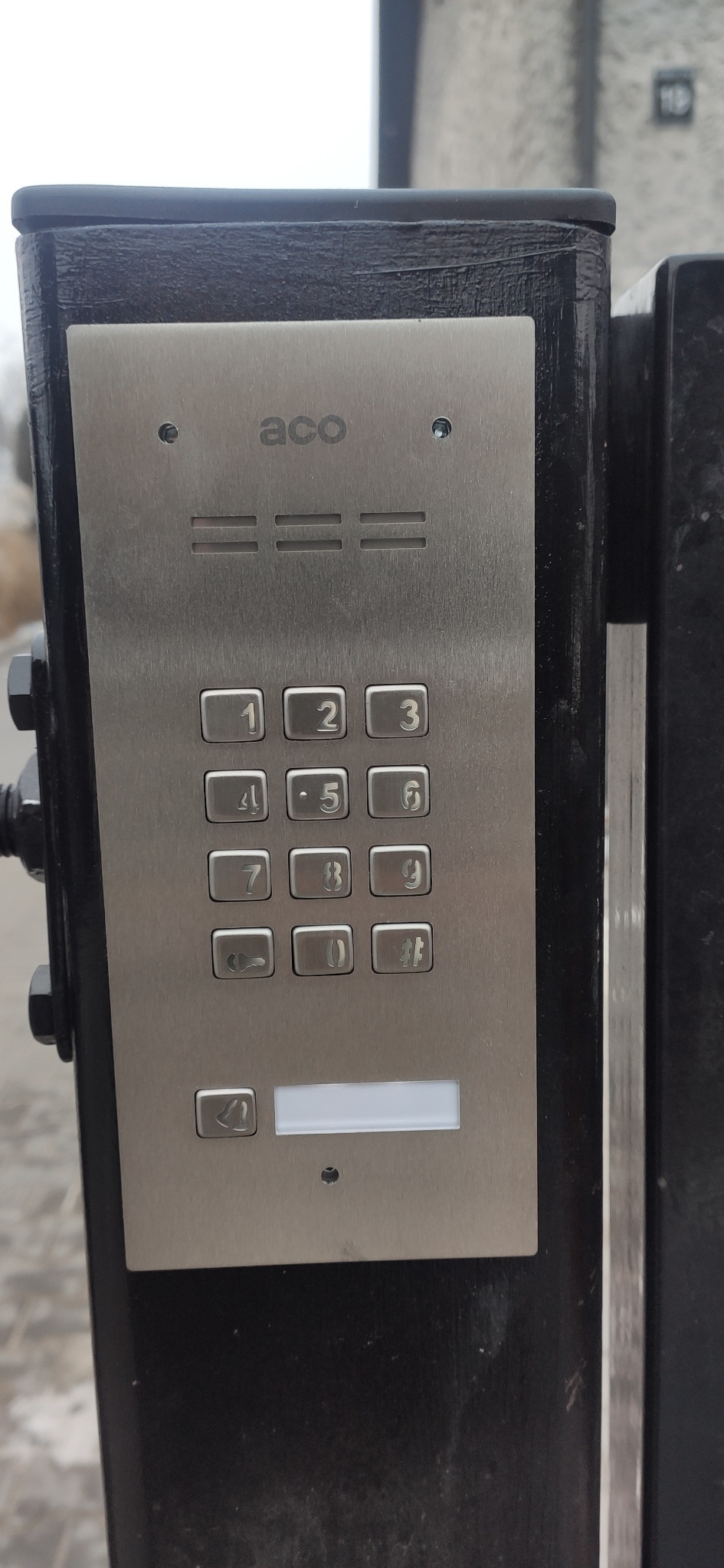 Metalowy panel domofonu z klawiaturą numeryczną i przyciskiem dzwonka, zamontowany na czarnej, metalowej ramie przy wejściu.