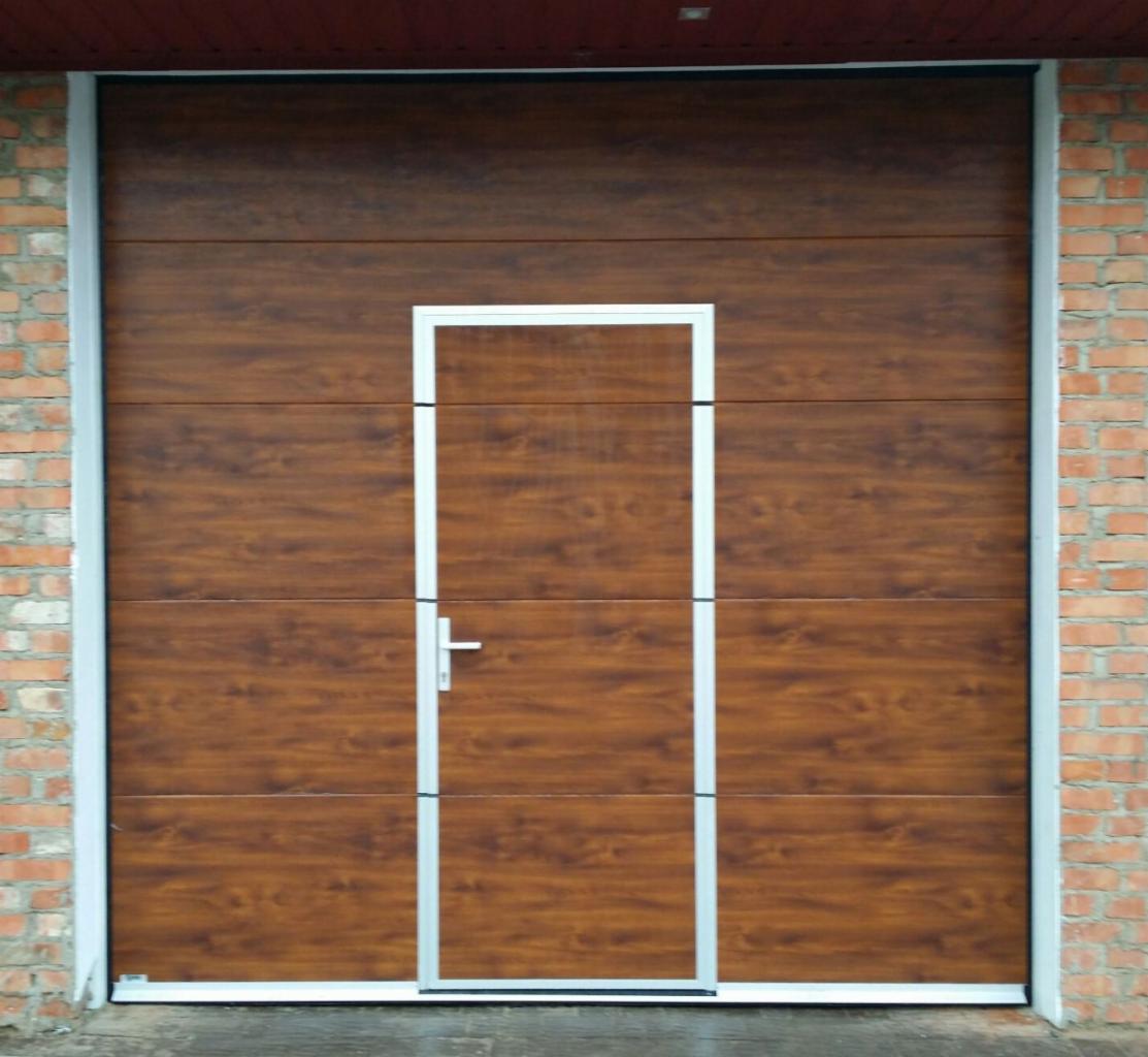 Segmentowa brama garażowa z drzwiami przejściowymi w kolorze drewna, z jasną ramą, zamontowana w murze z czerwonej cegły.