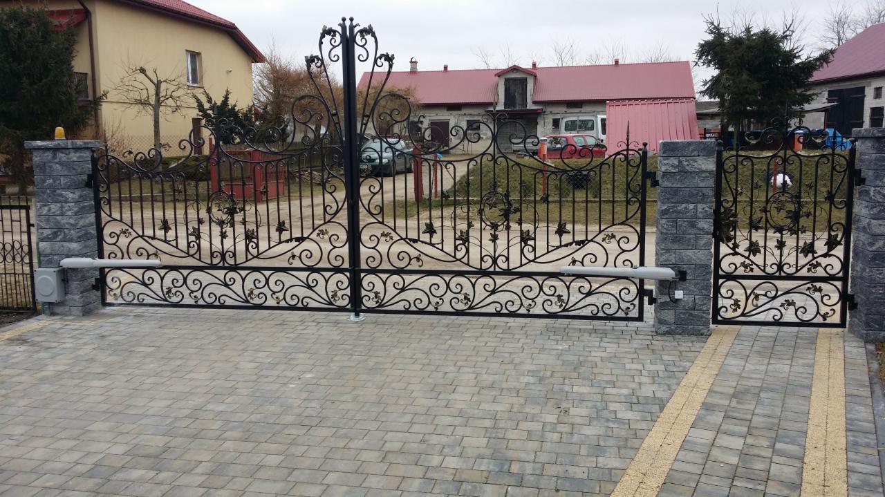Otwarta, dwuskrzydłowa brama wjazdowa z czarnego metalu o dekoracyjnych, roślinnych ornamentach, zamontowana na słupkach z szarej kostki, z widocznym napędem ramieniowym i kostką brukową...