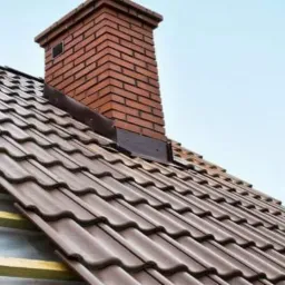 Fragment dachu z brązową dachówką ceramiczną i kominem z czerwonej cegły, widoczne elementy więźby dachowej w trakcie montażu.