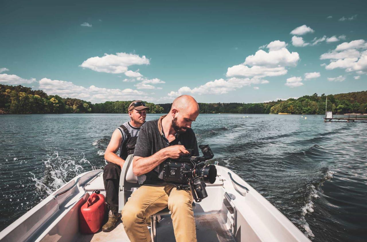 Operator kamery z brodą filmuje z łodzi na jeziorze, drugi mężczyzna siedzi obok, w tle zalesiony brzeg i błękitne niebo z chmurami.