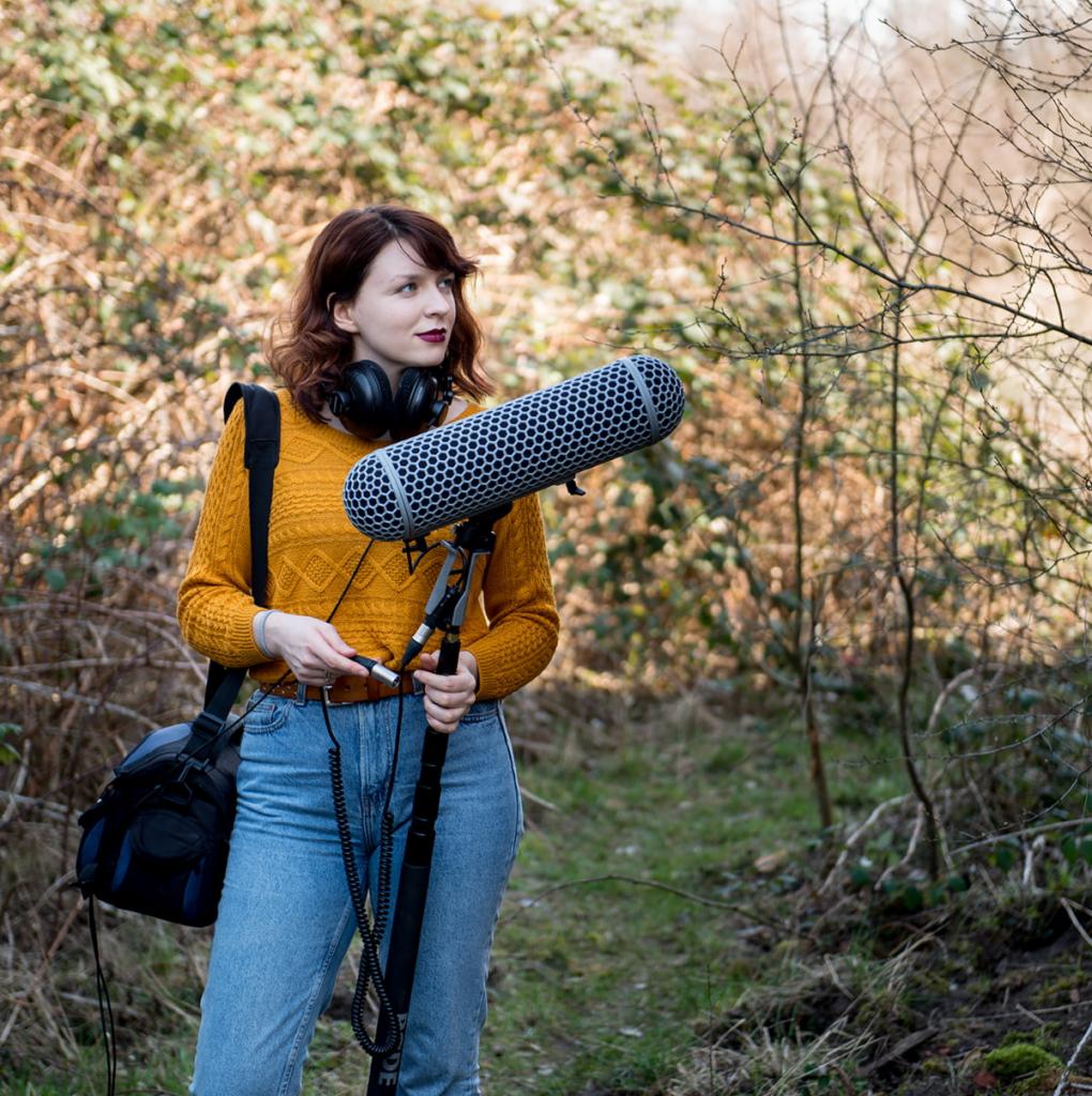 Kobieta z mikrofonem kierunkowym i słuchawkami na tle zarośli, prawdopodobnie podczas nagrywania dźwięku w plenerze.