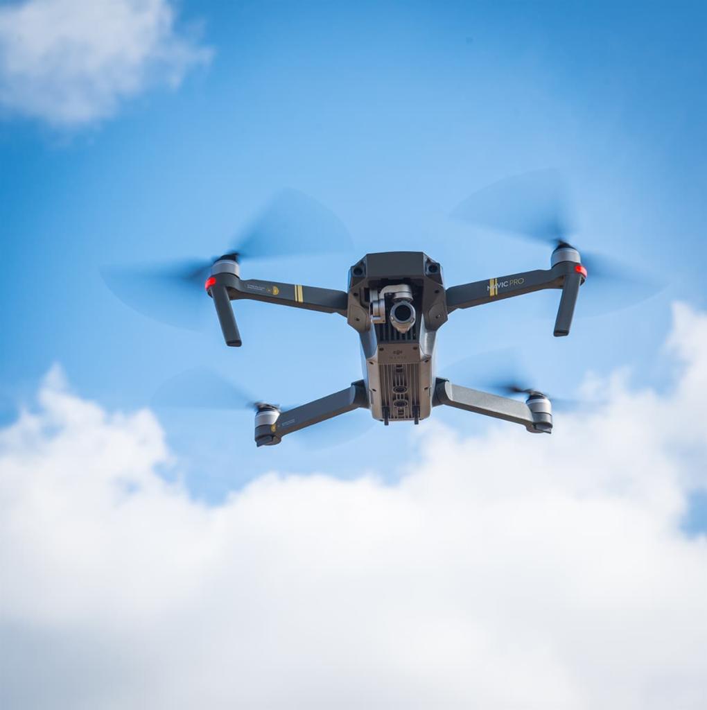 Szary dron Mavic Pro w locie na tle błękitnego nieba z białymi obłokami, widok od dołu, wirujące śmigła.