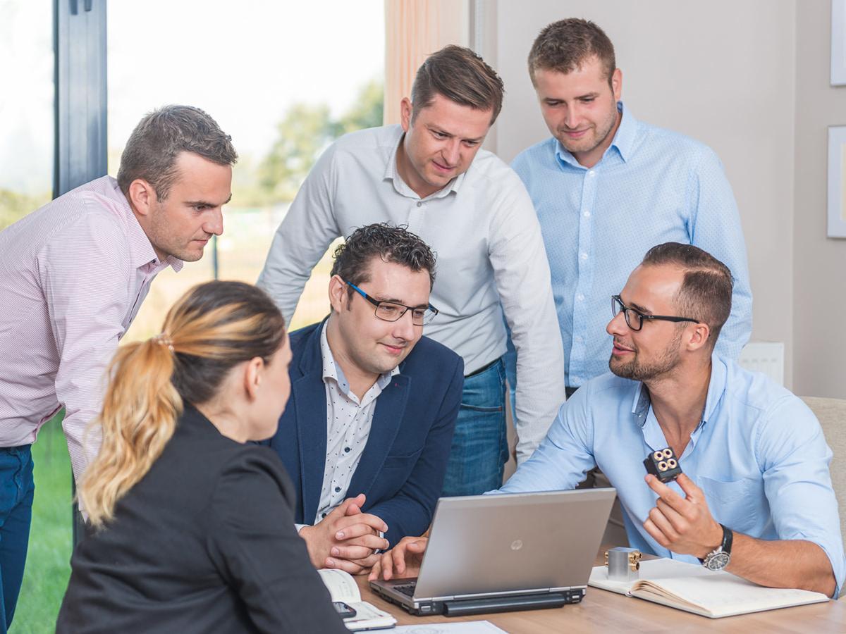 Zespół sześciu osób, w tym jedna kobieta, pochyla się nad laptopem na drewnianym stole, dyskutując o projekcie. Jeden z mężczyzn trzyma w ręku czarny element z ośmioma okrągłymi otworami.