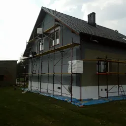 Dom jednorodzinny w trakcie prac związanych z izolacją termiczną elewacji, widoczne rusztowania, styropian oraz zabezpieczająca folia na trawniku.