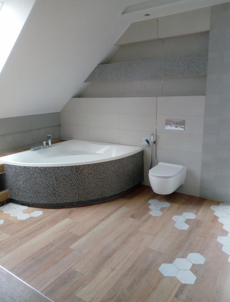 Nowoczesna łazienka na poddaszu z asymetryczną wanną obudowaną mozaiką, wiszącą toaletą i drewnopodobną podłogą z białymi heksagonalnymi wstawkami.