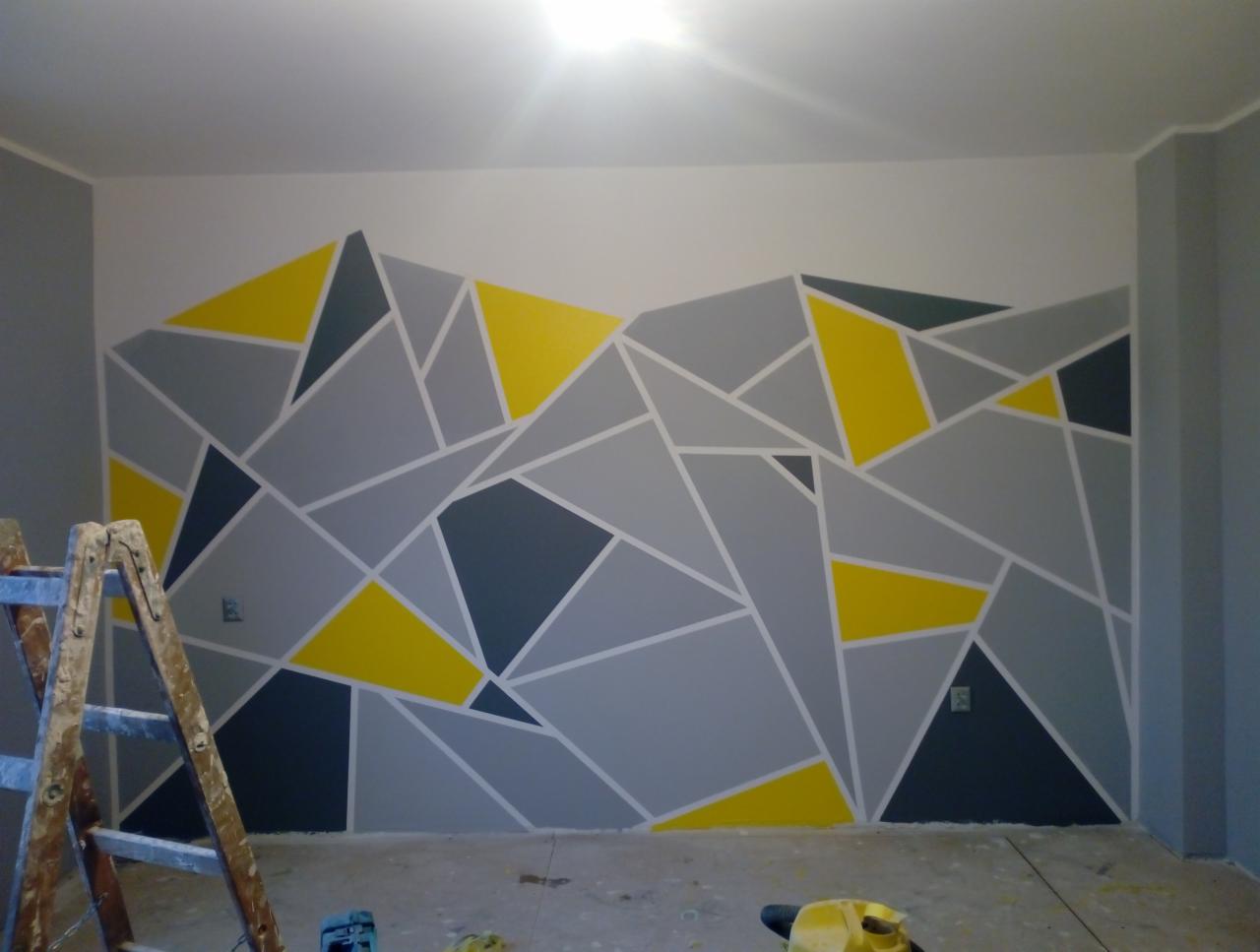 Ściana w pokoju z geometrycznym wzorem w odcieniach szarości i żółci, oddzielona taśmą malarską, widoczna drabina i narzędzia malarskie na podłodze.