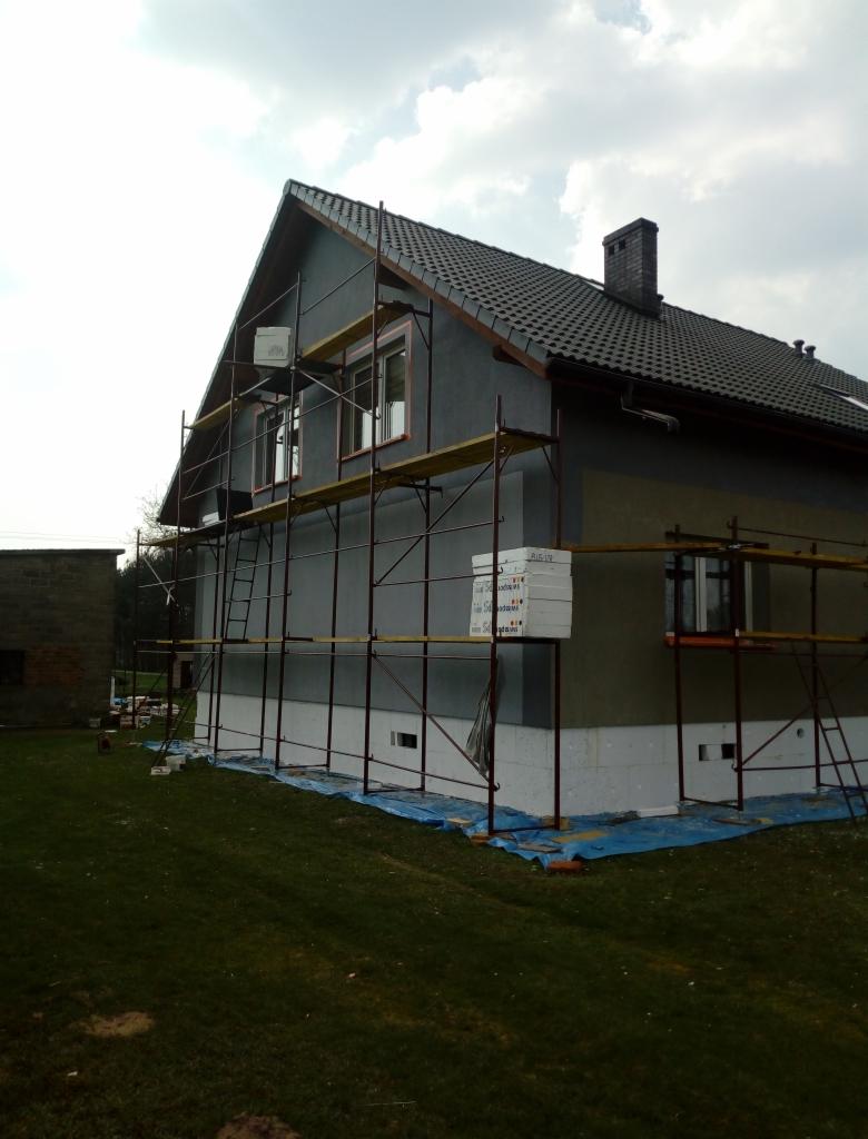 Dom jednorodzinny w trakcie prac związanych z izolacją termiczną elewacji, widoczne rusztowania, styropian oraz zabezpieczająca folia na trawniku.
