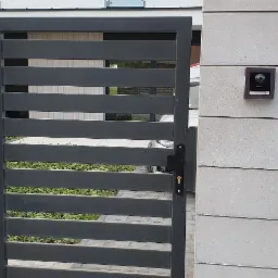 Zewnętrzny panel wideodomofonu IP zamontowany na słupku ogrodzeniowym z szarej cegły, widoczna furtka z poziomych profili i przew&oacute;d instalacyjny.