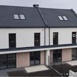 Nowoczesny bliźniak z systemem alarmowym: widoczna centrala alarmowa na elewacji, grafitowy dach z oknami dachowymi, duże przeszklenia na parterze.