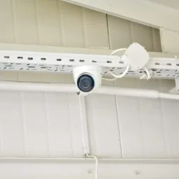 Biała kamera IP kopułkowa zamontowana na białej szynie kablowej pod sufitem, widoczne przewody i puszka instalacyjna.