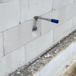 Szara rura wodna wystająca ze ściany z betonu komórkowego, z widocznymi złączkami i niebieską zaślepką na końcu, na tle surowego otoczenia budowlanego.