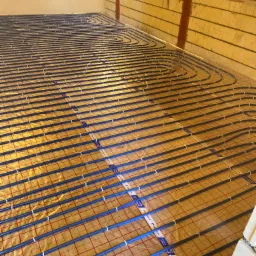 Rozłożone rury ogrzewania podłogowego na folii izolacyjnej w trakcie montażu w Zielonce, widoczne mocowania rur do folii i fragment ściany.