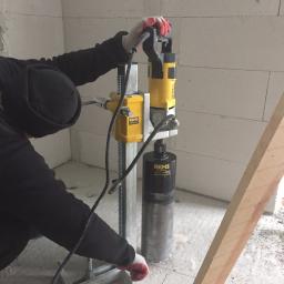 Profus - Hydraulik wierci otwór w ścianie z betonu komórkowego za pomocą wiertnicy diamentowej REMS Simplex 2, widoczne narzędzia i fragment drewnianej podpory