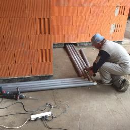 Profus - Instalator w trakcie łączenia rur w nowo wzniesionej ścianie z cegły ceramicznej. Na podłodze leżą dodatkowe rury i wiertarka z przedłużaczem.