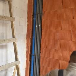Profus - Wewnętrzna instalacja rur wodnych i kanalizacyjnych w nowym budynku, widoczne rury szare i niebieskie, drabina drewniana, ściany z cegły ceramicznej porotherm i bloczków białych.