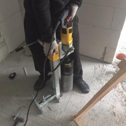 Profus - Pracownik w rękawicach wierci otwór w ścianie z betonu komórkowego za pomocą wiertnicy diamentowej REMS, widoczne narzędzia i fragment drewnianej konstrukcji.