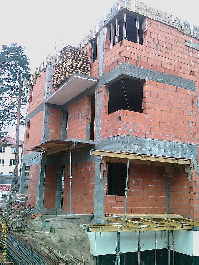 Budowa domu w stanie surowym otwartym: ściany z czerwonej cegły, częściowo zbrojone, z widocznymi otworami okiennymi i stertą palet na górnej kondygnacji, podpory szalunkowe i zbrojenie...