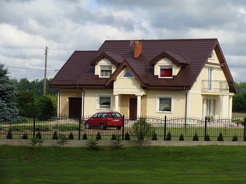 Żółty dom jednorodzinny z brązowym dachem z blachodachówki, balkonem z kutą balustradą i widocznym czerwonym samochodem osobowym za czarnym, kutym ogrodzeniem.