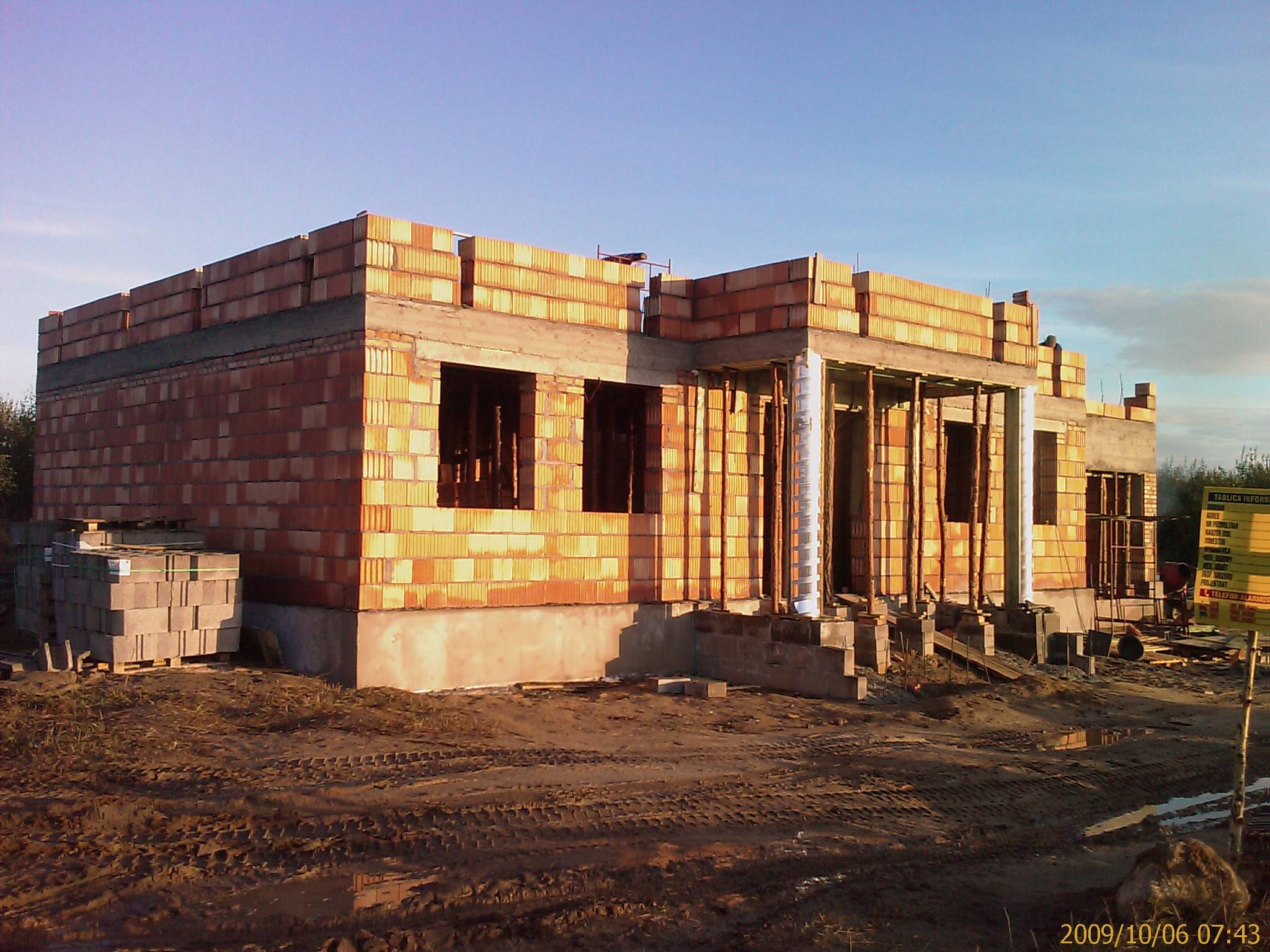 Niedokończony, jednopiętrowy budynek z czerwonej cegły, widoczny w świetle wczesnego poranka, z częściowo wzniesionymi ścianami i otworami okiennymi, na nierównym, błotnistym terenie budowy.