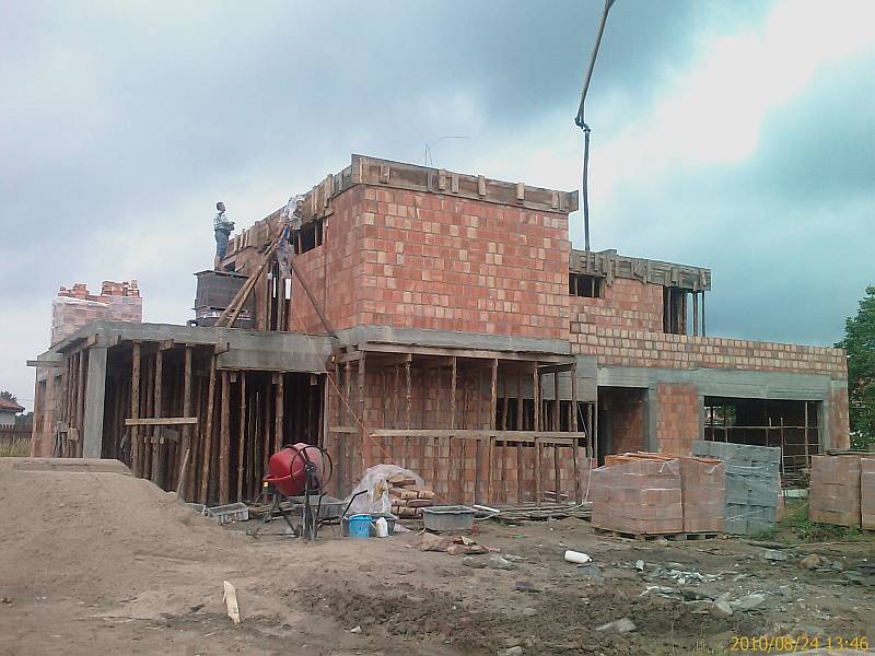 Budowa domu jednorodzinnego w stanie surowym otwartym: cegła ceramiczna, betonowy strop, szalunki drewniane, widoczny betonomieszarka i stos cegieł na placu budowy, pochmurne niebo w tle.