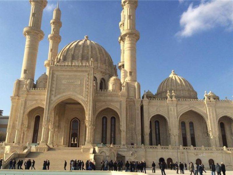 Monumentalna, jasna budowla z licznymi kolumnami i łukami, zwieńczona kopułami, z grupą ludzi u podnóża na tle błękitnego nieba.