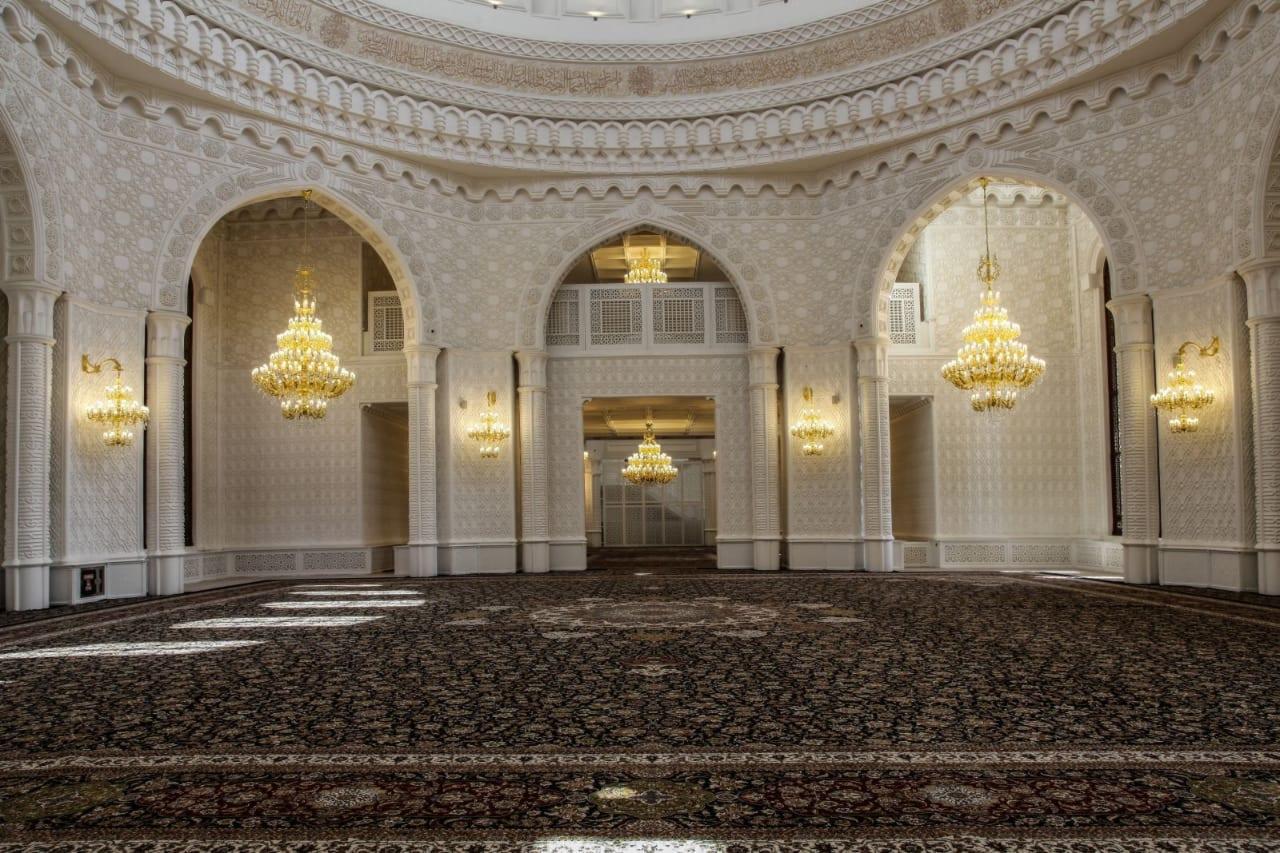 Przestronne wnętrze meczetu z białymi ścianami zdobionymi ornamentami, bogato zdobionym dywanem i licznymi kryształowymi żyrandolami, oświetlone naturalnym światłem wpadającym przez okna.