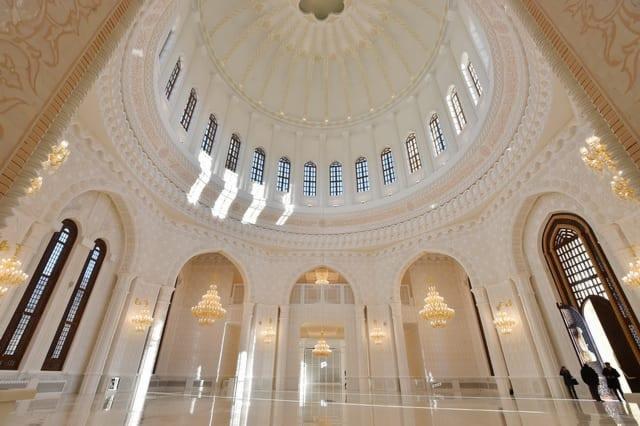 Jasne, przestronne wnętrze meczetu z kopułą, zdobieniami i żyrandolami; dwie osoby w oddali przy wejściu.