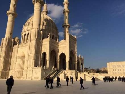 Widok na monumentalny, jasny budynek meczetu z czterema minaretami i kopułą na tle błękitnego nieba, z grupą ludzi spacerujących po placu przed budynkiem.