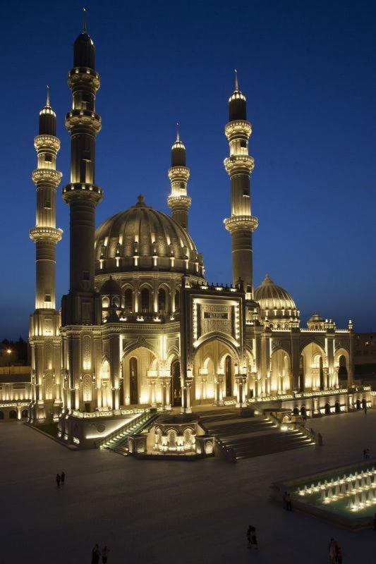 Oświetlona nocą monumentalna meczet z kopułą i minaretami, widoczne schody wejściowe i fontanna.