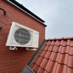 KRW SERWIS - Zewnętrzna jednostka klimatyzacji Mitsubishi zamontowana na ceglanej ścianie budynku, widoczna z perspektywy dachu pokrytego czerwoną dachówką.