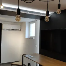 Wnętrze pokoju z zainstalowaną klimatyzacją Haier na ścianie, widoczne lampy wiszące z żarówkami Edisona, czarna ściana z telewizorem i regał, jasna ściana z oknem.