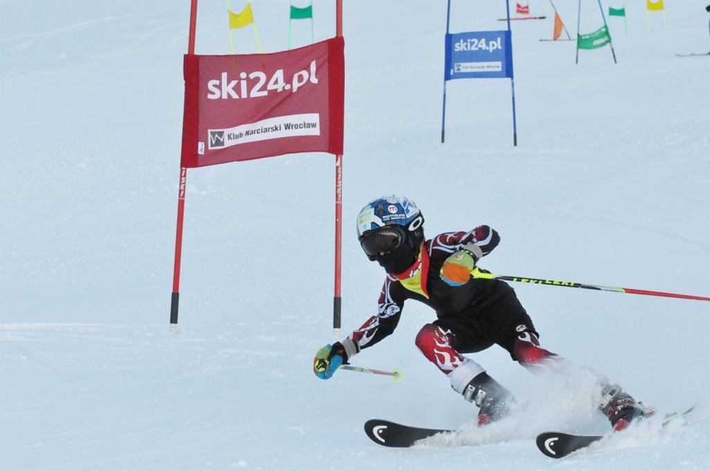 Młody narciarz w czarno-czerwonym kombinezonie podczas slalomu, sponsorowany przez ski24.pl, widoczny baner reklamowy ski24.pl i Klub Narciarski Wrocław.