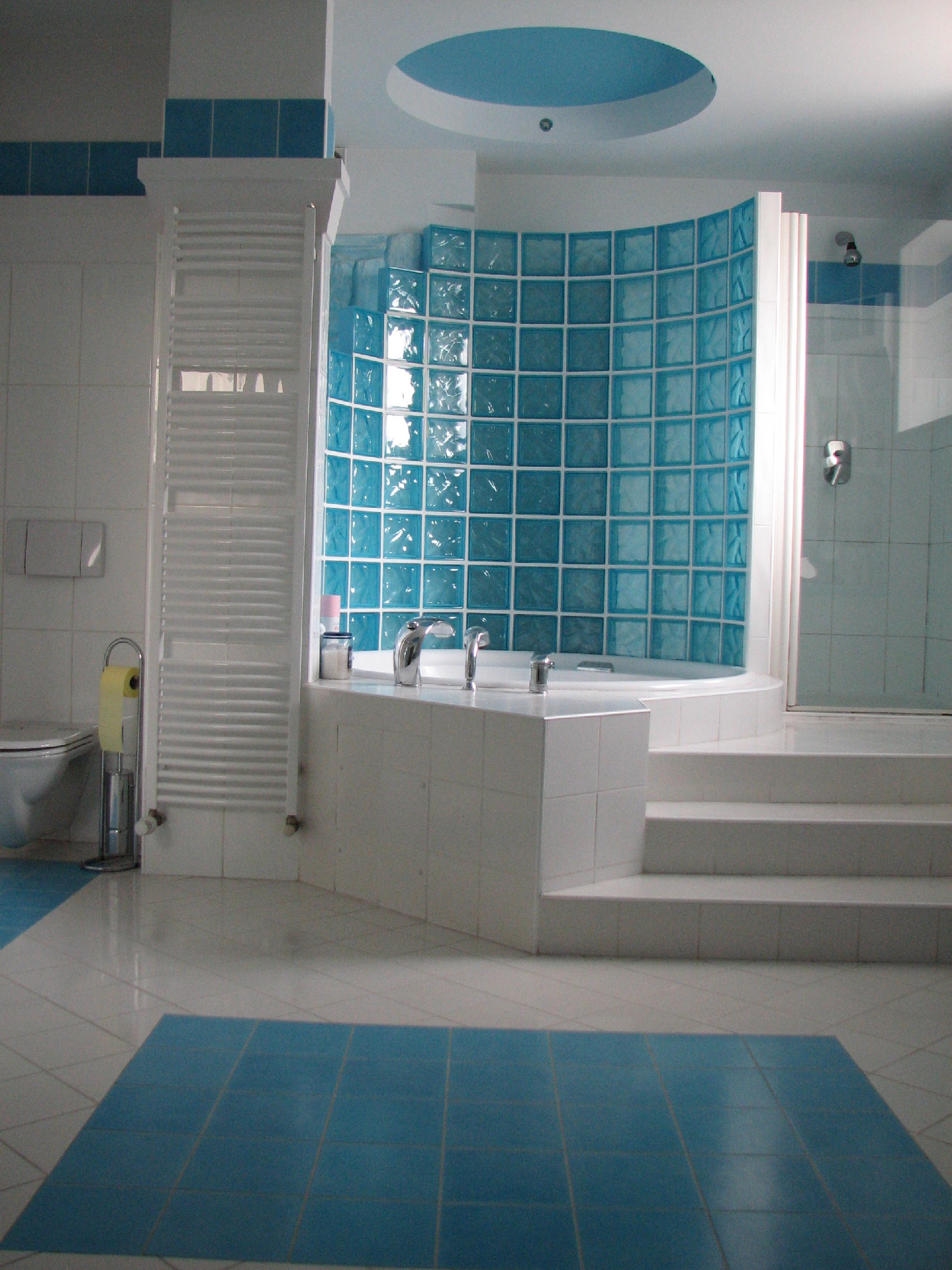 Biało-błękitna łazienka z wanną otoczoną ścianką z luksferów, widoczne schodki prowadzące do wanny oraz grzejnik drabinkowy.