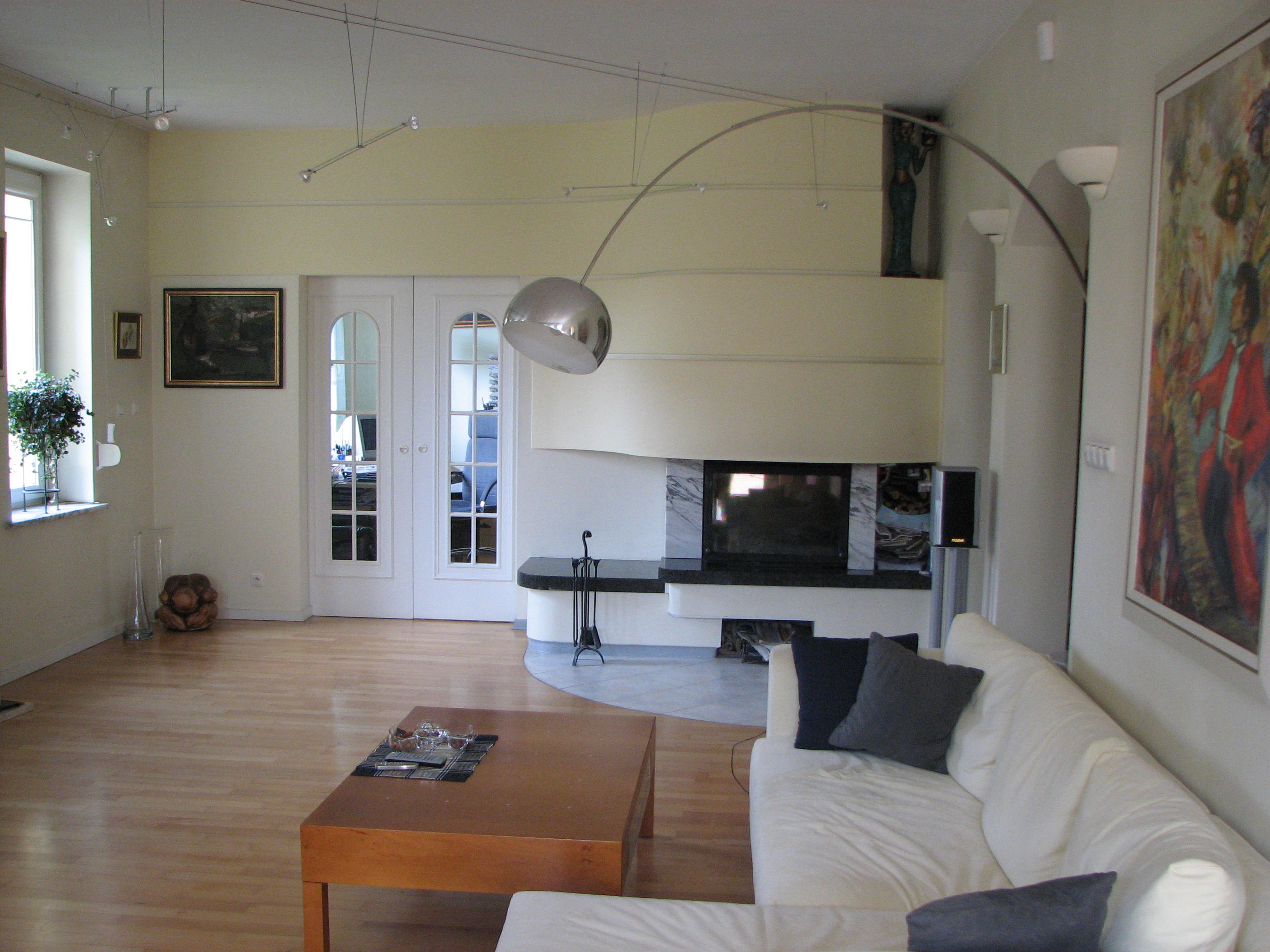 Jasny salon z drewnianą podłogą, białymi ścianami, kominkiem z marmurową obudową i nowoczesnym oświetleniem, widok na okno z rośliną i drzwi z przeszkleniami.