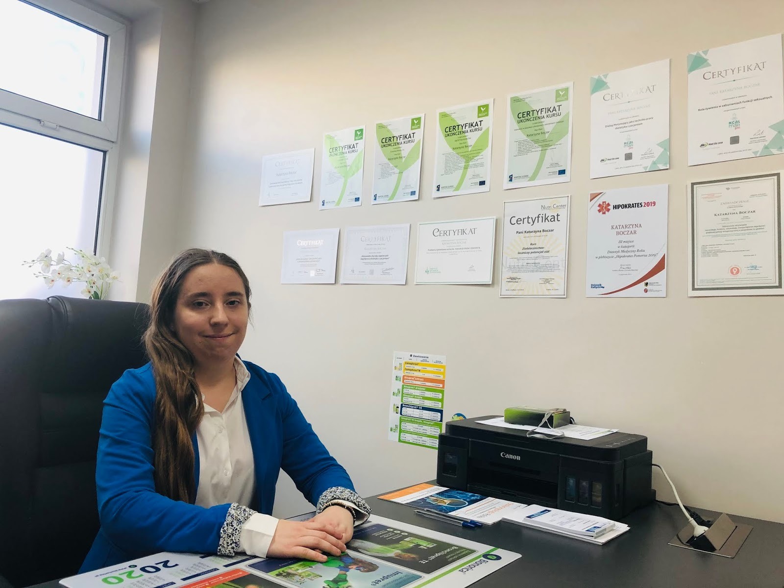 Kobieta w niebieskiej marynarce siedzi za biurkiem w biurze, na ścianie za nią wiszą certyfikaty i dyplomy, na biurku leżą materiały reklamowe i drukarka Canon.