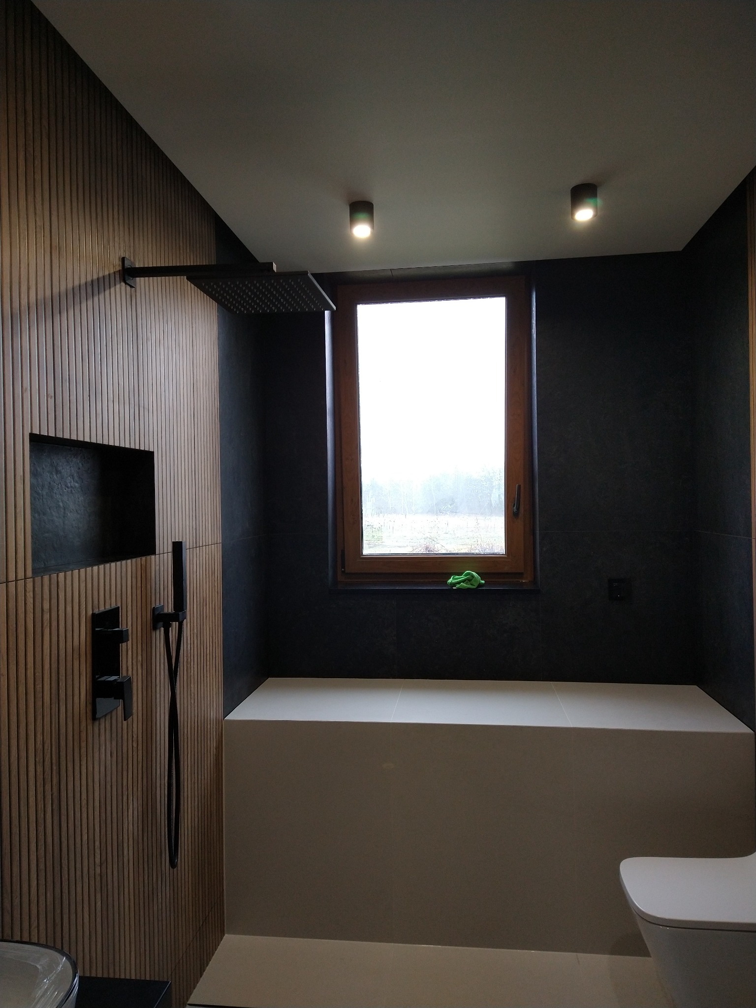 Nowoczesna łazienka z drewnianą ścianą z wnęką, czarną armaturą prysznicową, oknem z drewnianą ramą i zieloną ściereczką na parapecie oraz zabudowaną białą wnęką.