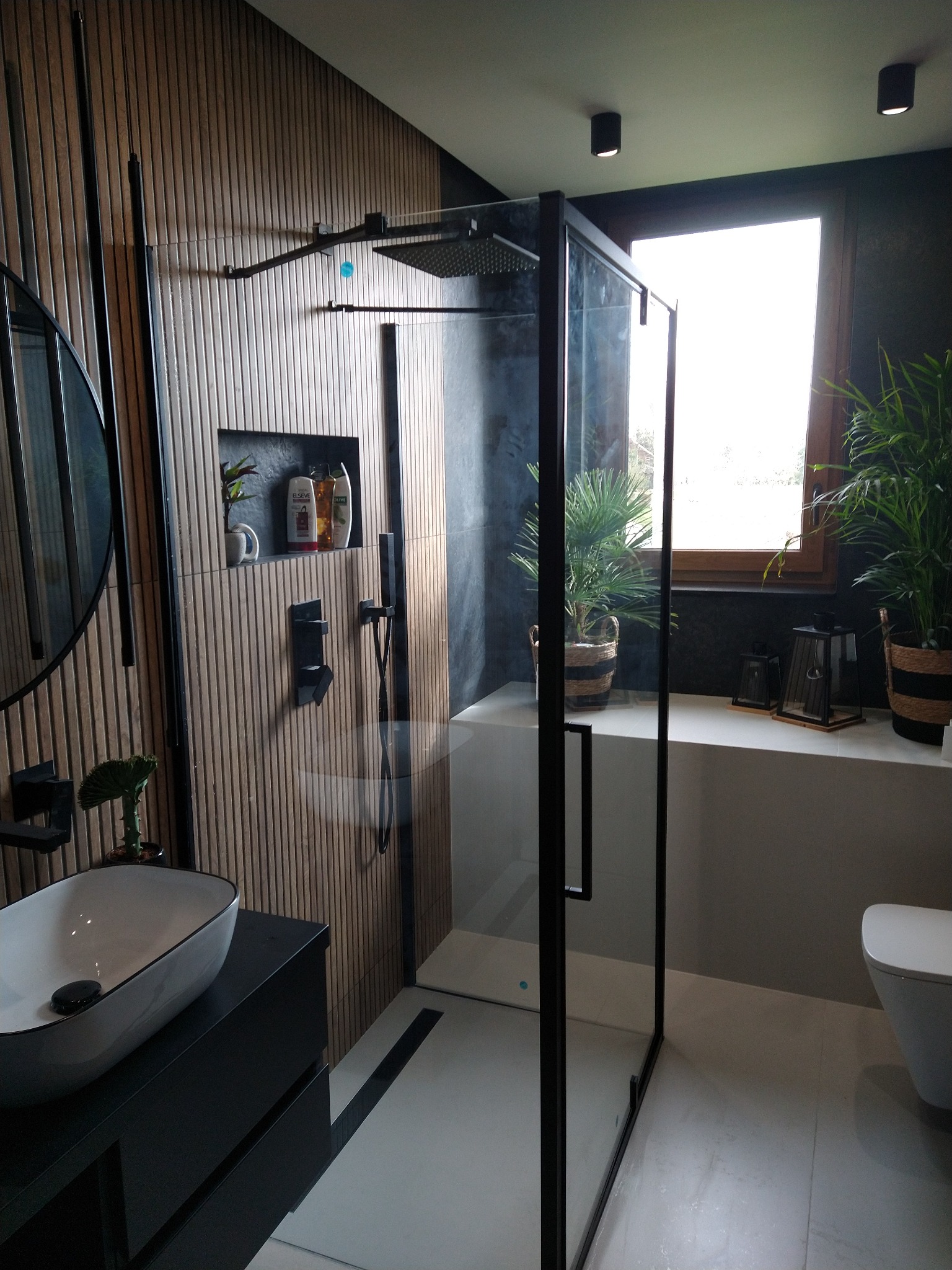 Nowoczesna łazienka z kabiną prysznicową typu walk-in z czarnymi profilami, umywalką nablatową na czarnej szafce, ścianą z drewnopodobnych paneli i roślinami doniczkowymi przy oknie.