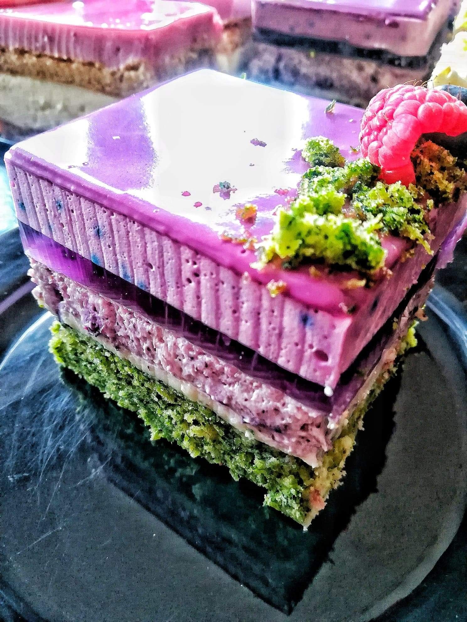 Kwadratowy deser warstwowy z zielonym spodem, różową masą i fioletową polewą lustrzaną, udekorowany zielonymi okruchami i maliną na błyszczącym czarnym talerzu.